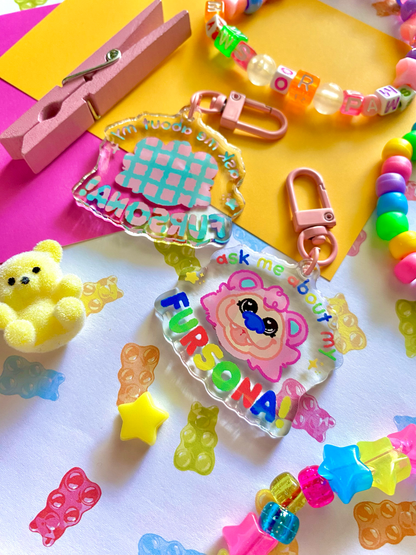 image features one keychains featuring original artwork by candy critters. 
details in the background of the product photo contains cute printed paper, colorful kandi bracelets, a teddy bear charm, and a pink clothes pin. (background items not included in purchase of keychain)

keychain is double sided.
left side features a drawing of the silhouette of design. 
right side features an image of a pink bear named caboose. surrounding text saying "ask me about my fursona"
hardware for keychain is pink