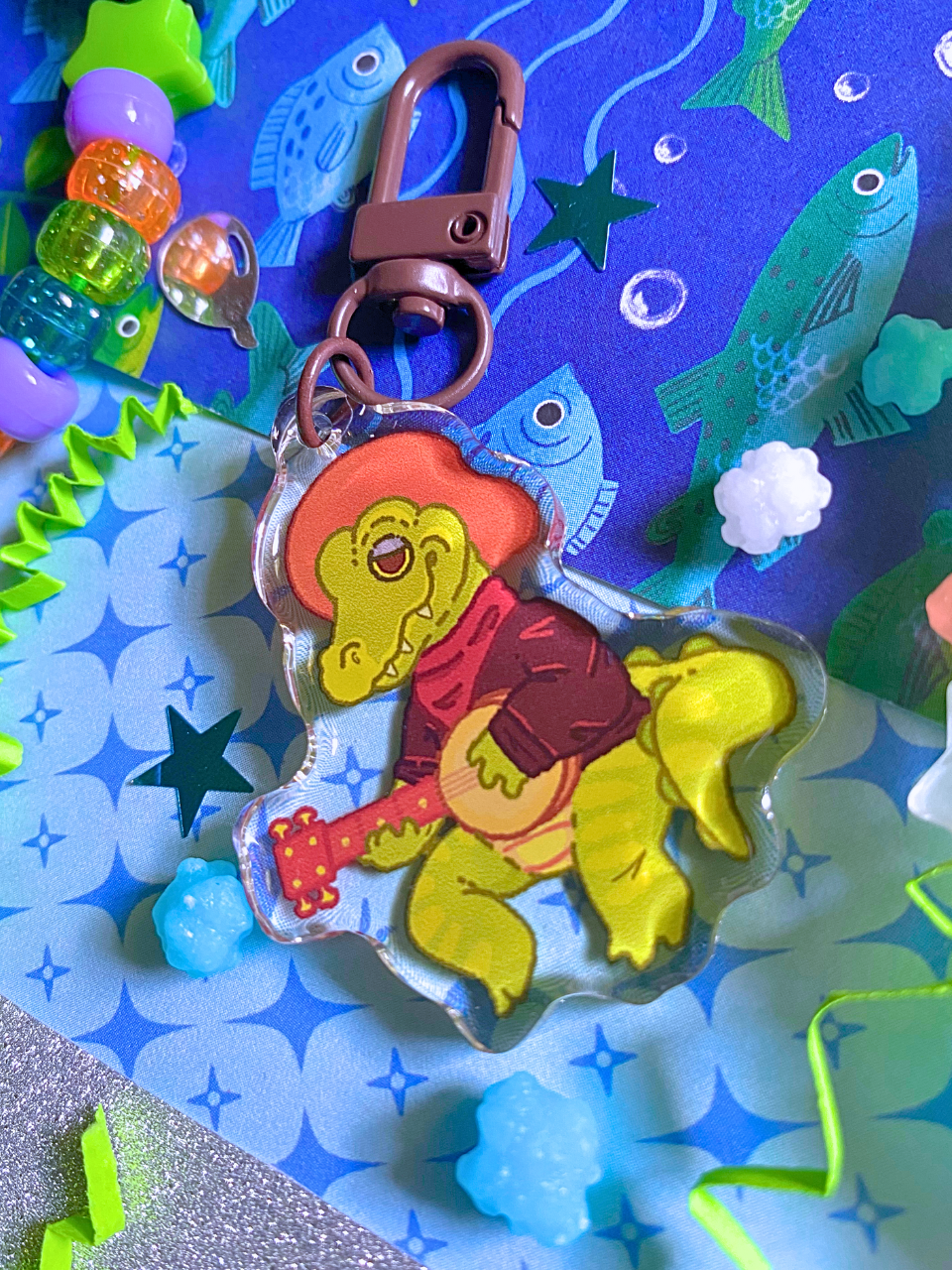 image features one keychain featuring original artwork by candy critters. 
details in the background of the product photo contains a collage of printed paper (fish, sparkles, and glitter), konpeito candy, confetti, and a kandi bracelet. (background items not included in purchase of keychain)

 keychain is double sided with same design on both sides. features a drawing of a green alligator with a hat, a shirt, and a banjo. the alligator is sitting playing banjo
the hardware for this keychain is black.