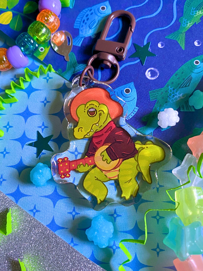 image features one keychain featuring original artwork by candy critters. 
details in the background of the product photo contains a collage of printed paper (fish, sparkles, and glitter), konpeito candy, confetti, and a kandi bracelet. (background items not included in purchase of keychain)

 keychain is double sided with same design on both sides. features a drawing of a green alligator with a hat, a shirt, and a banjo. the alligator is sitting playing banjo
the hardware for this keychain is black.