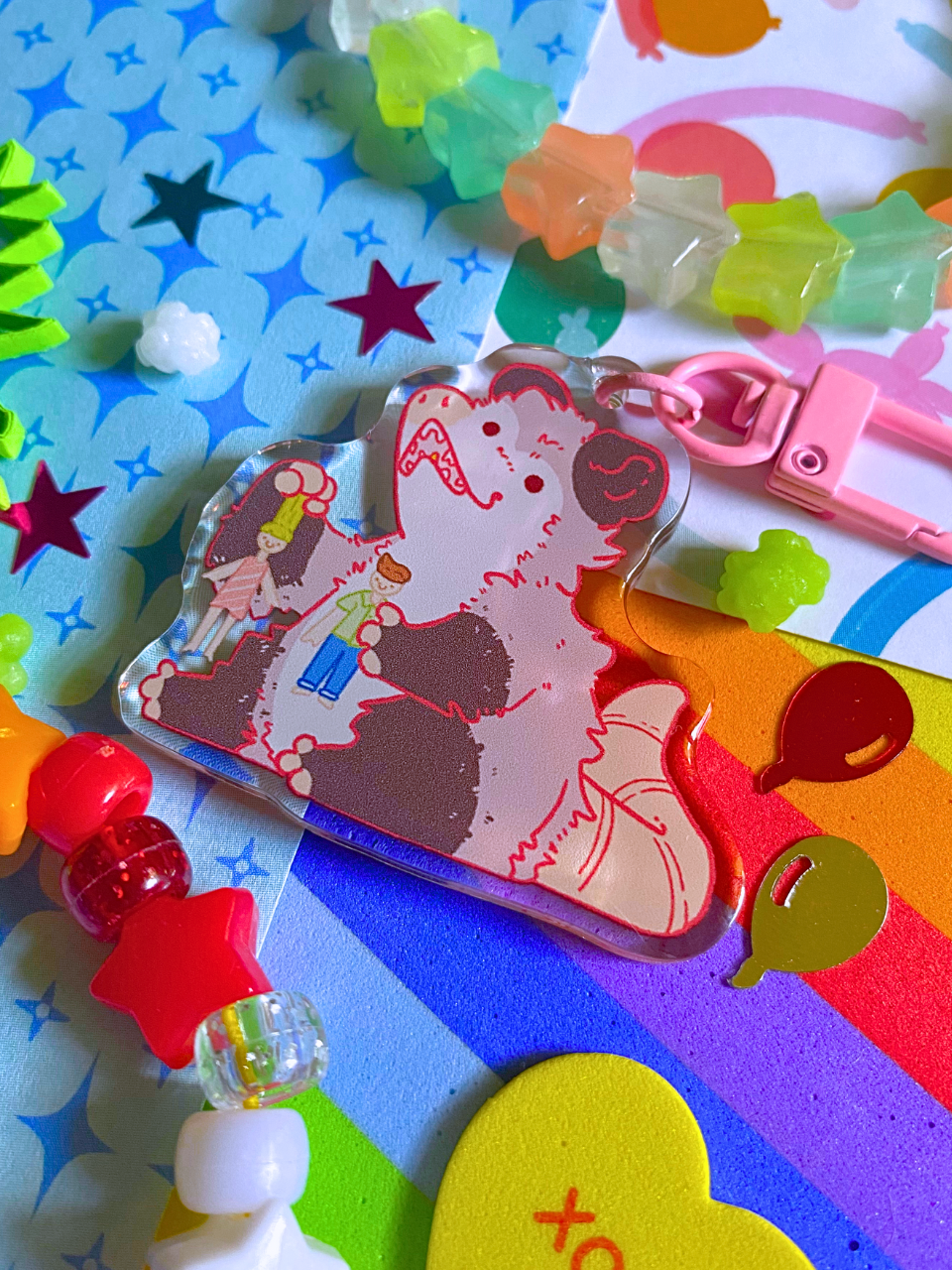 image features one keychain featuring original artwork by candy critters. 
details in the background of the product photo contains a collage of printed paper (balloon ani, sparkles, and rainbow), konpeito candy, confetti, and a kandi bracelet. (background items not included in purchase of keychain)

 keychain is double sided with same design on both sides. features a drawing of a possum playing with a girl and boy doll. 
the hardware for this keychain is pink.