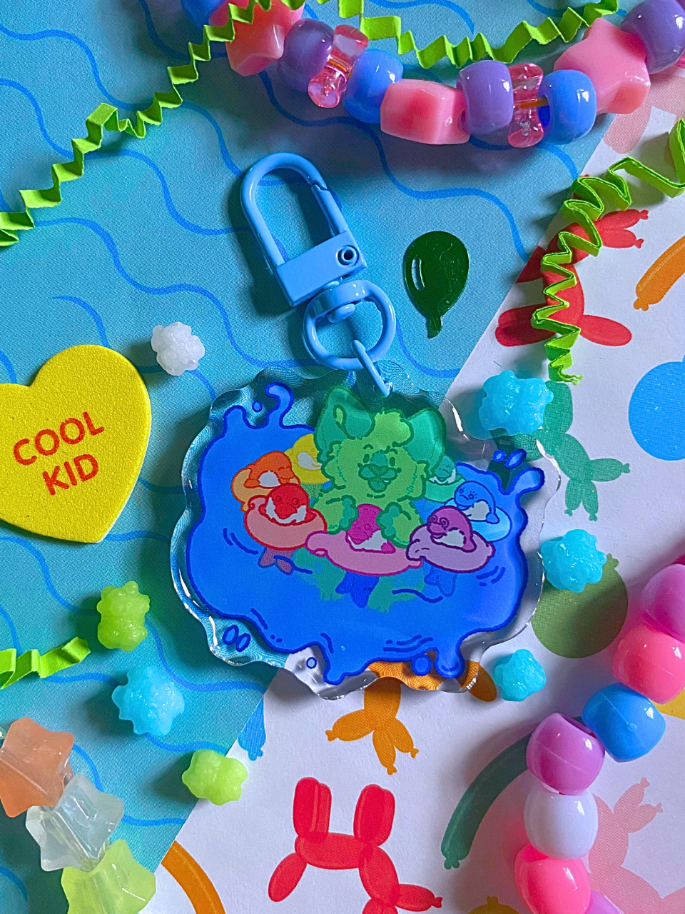 image features one keychain featuring original artwork by candy critters. 
details in the background of the product photo contains a collage of printed paper (waves, sparkles, and ballon animals), konpeito candy, confetti, and a kandi bracelet. (background items not included in purchase of keychain)

 keychain is double sided with same design on both sides. features a drawing of a green cat bathing in splashing water, playing with multicolor  floating dolphin toys
the hardware for this keychain is blue.