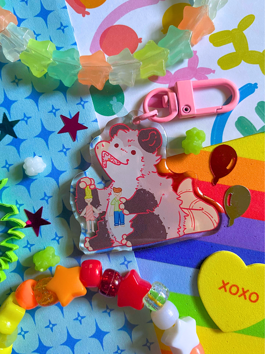 image features one keychain featuring original artwork by candy critters. 
details in the background of the product photo contains a collage of printed paper (balloon ani, sparkles, and rainbow), konpeito candy, confetti, and a kandi bracelet. (background items not included in purchase of keychain)

 keychain is double sided with same design on both sides. features a drawing of a possum playing with a girl and boy doll. 
the hardware for this keychain is pink.